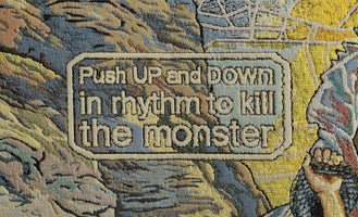 Ein gewebter Wandteppich in Nahaufnahme. In der Mitte steht in einem Kasten der Text: Push UP and DOWN in rhythm to kill the monster. Die Farben sind hauptsächlich Blau, Gelb und Braun.