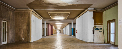 Langer, renovierungsbedürftiger Flur in Zentralperspektive mit einem gefliesten Boden, einer beschädigten Decke und unfertigen Wänden. An der rechten Wand lehnt ein Schild mit der Aufschrift MMM 1989.
