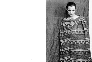 Ein Schwarz-Weiß-Foto zeigt eine Frau mit blasser Haut, zurückgekämmtem dunklen Haar und dramatischem, spitzem Eyeliner. Sie trägt einen voluminösen, gewebten Umhang mit einem geometrischen Muster und blickt mit intensivem Ausdruck direkt in die Kamera, vor einem dunklen, zerknitterten Hintergrund.