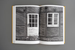 Ein aufgeschlagenes Buch mit gelbem Einband liegt auf einer grauen Oberfläche und zeigt eine Schwarz-Weiß-Fotografie eines alten Fachwerkhauses mit einer verwitterten weißen Holztür und einem Fenster.