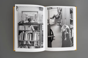 Ein aufgeschlagenes Buch mit gelbem Einband liegt auf einer grauen Oberfläche und zeigt eine Doppelseite mit Schwarz-Weiß-Fotografien. Die Fotos bilden einen vollgestellten Raum ab, mit einem Bücherregal, einer Büste, Bildern, einer kopfüber hängenden Figur und einem Papierskelett eines Dinosauriers.