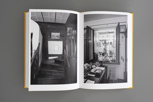 Ein aufgeschlagenes Buch mit gelbem Einband zeigt zwei Schwarz-Weiß-Fotografien. Links ist eine dunkle Holztreppe in einem engen Treppenhaus zu sehen, rechts ein vollgestellter Schreibtisch vor einem offenen Fenster mit Blick in einen weiteren Raum.