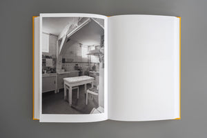 Ein aufgeschlagenes Buch mit gelbem Einband liegt auf einer grauen Oberfläche. Die linke Seite zeigt eine Schwarz-Weiß-Fotografie einer altmodischen Küche, die rechte Seite ist leer.