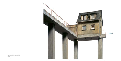 Ein kleines, verwittertes Haus steht auf hohen, dicken Betonpfeilern. Ein langer Steg mit einem rostigen Geländer führt auf der gleichen Ebene zum Haus. Die gesamte Konstruktion ist ein Modell vor einem weißen Hintergrund.