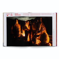 Ein aufgeschlagenes Buch zeigt eine Fotografie, die eine Gruppe von Menschen bei Nacht in einer hügeligen Landschaft darstellt. Die Personen tragen durchsichtige Plastikponchos und sind eng um ein Feuer versammelt, das sie in ein warmes, orangefarbenes Licht taucht. Der darüber stehende Text lautet Einzelfotos John Moore und Nächtliche Grenzüberschreitung.