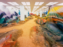 Eine raumfüllende Kunstinstallation in einem großen Raum mit weißen Säulen und einer hell erleuchteten Rasterdecke. Ein sandiger Pfad führt durch eine Landschaft aus Erdhügeln und riesigen, bunt bemalten Stoffbahnen, die die Wände verkleiden.