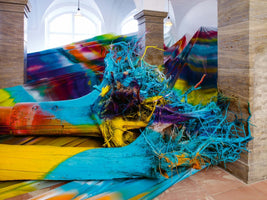 Eine große, farbenfrohe Kunstinstallation in einem hellen Raum mit Steinsäulen. Mehrere große Baumstämme und deren verfilzte Wurzeln sind mit leuchtend blauer, gelber und violetter Farbe besprüht und liegen auf einer ebenfalls bunten, drapierten Stoffbahn.