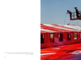 Eine Buchdoppelseite, die rechts ein Foto von zwei Personen auf einer Hebebühne zeigt, die ein Gebäude und den Boden mit leuchtend roter Farbe besprühen, wobei das Dach des Gebäudes rote und weiße Streifen aufweist, alles unter einem klaren blauen Himmel.