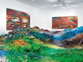 Eine helle Galerie mit einer Bodeninstallation aus bunten Pigmenthaufen in Grün, Blau und Orange, die eine Art Tal bilden. An den weißen Wänden hängen zwei große, farbenfrohe, abstrakte Gemälde.