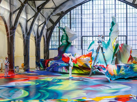 Zwei Künstler in weißen Schutzanzügen arbeiten in einer großen Halle mit gotischen Säulen an einem riesigen, abstrakten Kunstwerk. Der gesamte Boden und mehrere große, skulpturale Formen sind mit lebhaften, wirbelnden Schichten aus roter, blauer, gelber und grüner Farbe bedeckt. Einer der Künstler steht auf einer Leiter und sprüht Farbe, während die Säulen zum Schutz in Plastik eingewickelt sind.