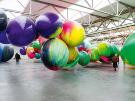 Innenansicht einer Kunstinstallation in einer großen Halle mit vielen riesigen, bunten, kugelförmigen Ballons, die in der Luft schweben und auf dem Boden liegen. Einige Besucher gehen zwischen den Kugeln umher.