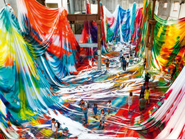 Eine Kunstinstallation aus der Vogelperspektive in einer großen Halle, in der Menschen zwischen riesigen, von der Decke hängenden und am Boden ausgebreiteten Stoffbahnen in leuchtenden Batik-Farben spazieren.