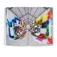 Ein aufgeschlagenes Buch zeigt eine raumfüllende Kunstinstallation in einer großen Halle. Lange, farbenfroh und abstrakt bemalte Stoffbahnen hängen von der Decke bis zum Boden und bilden einen langen Korridor, in dem eine winzige Person steht.