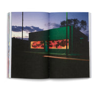 Ein aufgeschlagenes Buch zeigt eine Fotografie eines leuchtend grünen Gebäudes an einer Straßenecke bei Dämmerung. Die Fenster sind von innen rot-orange erleuchtet und mit Graffiti versehen, während Lichtspuren eines Autos über die dunkle Straße im Vordergrund ziehen.