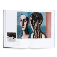 Ein aufgeschlagenes Kunstbuch zeigt ein surrealistisches Gemälde über eine Doppelseite. Es sind zwei Büsten vor einem Meer zu sehen. Die linke Büste hat ein fragmentiertes Gesicht, durch das der Himmel sichtbar wird. Die rechte Büste ist aufgebrochen und gibt den Blick auf ein dunkles Inneres mit vertikalen Strängen und Kugeln frei. Links unten auf der Seite befindet sich ein kleines Schwarz-Weiß-Foto.