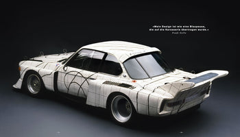 Ein weißer BMW 3.0 CSL Rennwagen, dessen Karosserie mit einem schwarz-weißen Gittermuster überzogen ist, das an eine Blaupause erinnert. Das von Frank Stella gestaltete Art Car wird schräg von hinten vor einem dunklen Hintergrund gezeigt.