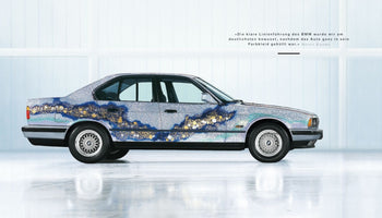 Seitenansicht des BMW Art Cars von Matazo Kayama in einem hellen Studio. Das Auto hat eine silberne Glitzerlackierung mit einem kunstvollen, abstrakten Muster in Blau-, Türkis- und Goldtönen entlang der Seite.