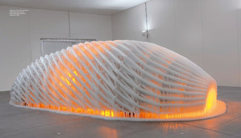 Eine große, kuppelförmige Skulptur in einem weißen Raum, deren gitterartige Struktur mit Eis bedeckt ist. Ein warmes, orangefarbenes Licht leuchtet aus dem Inneren und scheint durch die eisige Hülle.
