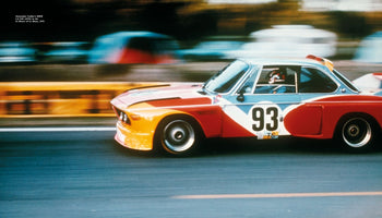 Eine Bewegungsaufnahme des bunten BMW 3.0 CSL Art Car von Alexander Calder mit der Nummer 93 auf einer Rennstrecke. Der Hintergrund ist durch die hohe Geschwindigkeit des Wagens verschwommen.