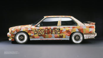 Ein BMW E30 M3 in der Seitenansicht, dessen Karosserie vollständig mit einem Kunstwerk von Michael Jagamara Nelson im Stil der Papunya-Tula-Malerei gestaltet ist. Das Design besteht aus unzähligen Punkten in erdigen Farben, die komplexe Muster und Symbole bilden, vor einem dunklen Hintergrund.