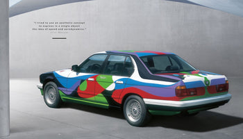 Ein BMW Art Car von César Manrique mit einer bunten, abstrakten Lackierung aus fließenden Formen in Blau, Rot, Grün und Lila, geparkt vor einer grauen Betonwand. Oben links steht ein Zitat des Künstlers.