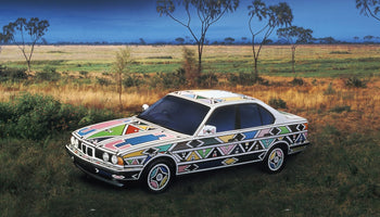 Ein BMW, kunstvoll bemalt mit bunten, geometrischen Mustern im Ndebele-Stil, steht in einer weiten, grasbewachsenen Savannenlandschaft unter einem dunkelblauen Himmel.