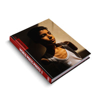 Das Buch World Press Photo 2025 liegt schräg auf einer weißen Oberfläche. Auf dem Cover befindet sich das Porträt eines Jungen in einem weißen Unterhemd, dessen Gesicht und Oberkörper von starkem Licht beleuchtet werden, was dramatische Schatten erzeugt.