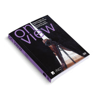 Ein Buch mit dem Titel on view liegt schräg auf einer weißen Oberfläche. Der Einband ist schwarz mit lila und weißer Schrift. Das Coverfoto zeigt die Spiegelung einer Person, die eine Kamera auf einem Stativ hält.