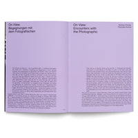 Ein aufgeschlagenes Buch, das auf einer weißen Oberfläche liegt. Die linke Seite hat einen lavendelfarbenen Kopfbereich mit dem Titel On View. Begegnungen mit dem Fotografischen, gefolgt von einem Textblock auf Deutsch. Die rechte Seite hat einen passenden Kopfbereich mit dem englischen Titel On View: Encounters with the Photographic, gefolgt von einem Textblock auf Englisch.
