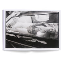 Schwarz-Weiß-Foto in einem aufgeschlagenen Buch: Eine weibliche Schaufensterpuppe liegt nackt mit einer Perlenkette in einer Glasvitrine. Sie blickt nach oben, neben ihrer Schulter liegt eine leuchtende Kugel.