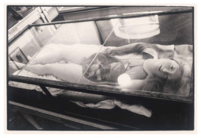 Eine Schwarz-Weiß-Aufnahme eines anatomischen Wachsmodells einer Frau, das auf dem Rücken in einer Glasvitrine liegt. Ihr Bauch- und Brustbereich ist geöffnet, um die inneren Organe und einen Fötus zu zeigen. Die Figur hat langes, helles Haar, einen gelassenen Gesichtsausdruck und trägt eine Perlenkette.