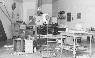 Eine Schwarz-Weiß-Aufnahme eines unordentlichen Künstlerateliers, gefüllt mit einer Staffelei, Tischen voller Kunstzubehör, einem Globus, Holzkisten und auf dem Boden verstreuten Papieren und Materialien.