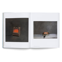 Ein aufgeschlagenes Buch auf einer weißen Fläche. Auf der linken Seite befindet sich eine Fotografie eines altmodischen, kastenförmigen Heizstrahlers mit leuchtend orangen Heizstäben vor einem dunklen Hintergrund. Auf der rechten Seite ist eine Fotografie eines alten, runden Waffeleisens auf einer hellgrauen Fläche, ebenfalls vor einem dunklen Hintergrund.