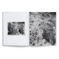 Ein aufgeschlagenes Buch auf weißem Hintergrund, das auf beiden Seiten körnige Schwarz-Weiß-Fotografien einer abstrakten, felsigen Textur zeigt. Links ist das Foto kleiner und gerahmt, rechts füllt es die gesamte Seite als extreme Nahaufnahme.
