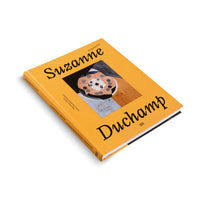 Ein gelbes Buch mit dem Titel Suzanne Duchamp Retrospektive liegt schräg auf einem weißen Hintergrund. Auf dem Umschlag befindet sich ein abstraktes Gemälde mit geometrischen Formen in der Mitte.