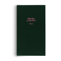 Ein Buch mit einem dunkelgrünen Stoffeinband, auf dem in rosafarbener Schrift HILMA af KLINT und darunter Flora steht.