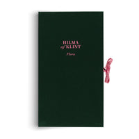 Ein tiefgrünes Buch mit dem Titel HILMA af KLINT Flora in rosa Schrift und einem rosa Schleifenband als Verschluss.