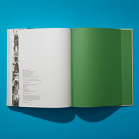 Von oben fotografiert liegt ein aufgeschlagenes Buch auf einer leuchtend blauen Fläche. Die linke Seite ist weiß und zeigt das Impressum, daneben eine schmale Spalte mit Schwarz-Weiß-Fotos. Die rechte Seite ist eine einfarbige, grüne Seite, gefolgt von weiteren grünen Seiten.
