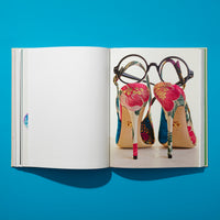 Ein aufgeschlagenes Buch liegt auf einer leuchtend blauen Oberfläche. Die linke Seite ist leer, während die rechte Seite ein Foto der Rückseite eines Paares bunter, geblümter High Heels zeigt, auf denen eine schwarze, runde Brille balanciert.