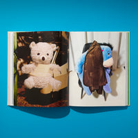 Ein aufgeschlagenes Fotobuch liegt auf einer leuchtend blauen Oberfläche. Die linke Seite zeigt einen hellen Teddybären, der mit Pappe umwickelt in einem Auto sitzt. Die rechte Seite zeigt ein blaues I-Aah-Stofftier auf einem Stuhl, über das eine braune Jacke gehängt ist.