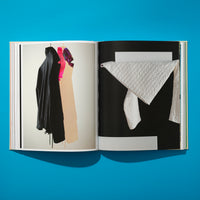 Eine Draufsicht auf ein aufgeschlagenes Buch, das auf einer leuchtend blauen Fläche liegt. Die linke Seite zeigt ein Foto von hängenden Kleidungsstücken, darunter eine schwarze Jacke. Die rechte Seite zeigt eine abstrakte Komposition mit einem weißen, gesteppten Stoff, der auf einem schwarz-weißen geometrischen Hintergrund drapiert ist.
