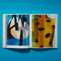 Ein aufgeschlagenes Buch liegt auf einer leuchtend blauen Oberfläche und zeigt zwei Modefotografien. Links ist ein Hemdkragen mit schwarzen Krawatten vor einem großen, hellen Kreis zu sehen. Rechts ist ein farbenfrohes, gestreiftes Strickoberteil von Prada mit großen, ovalen Löchern abgebildet.
