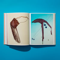 Ein aufgeschlagenes Buch auf einer leuchtend blauen Oberfläche, das zwei Fotos zeigt: links eine braune Ledertasche mit Fransen und rechts ein kariertes Tuch, das über Seilbahnkabeln mit zwei Gondeln drapiert ist.