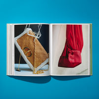 Ein aufgeschlagenes Buch liegt auf einer leuchtend blauen Oberfläche, das von oben fotografiert wurde. Auf der linken Seite des Buches ist eine braune, gesteppte Handtasche mit weißem Shearling-Besatz und einem goldenen Kettengriff abgebildet. Auf der rechten Seite ist eine Nahaufnahme eines leuchtend roten Blusenärmels zu sehen.