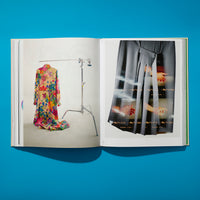 Eine Draufsicht auf ein aufgeschlagenes Buch, das auf einer leuchtend blauen Fläche liegt. Auf der linken Seite hängt ein buntes Blumenkleid an einem Kleiderständer. Auf der rechten Seite ist ein grauer Rock abgebildet, über den transparente Filmstreifen mit Bildern von Händen gelegt sind.