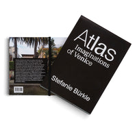 Ein schwarzes Buch mit dem weißen Titel Atlas Imaginations of Venice von Stefanie Bürkle. Das Buch liegt auf einer weißen Fläche, wobei das Cover die Rückseite teilweise verdeckt, auf der ein Foto von Palmen und einem Gebäude zu sehen ist.